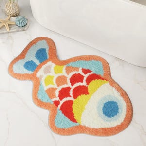 May include: A fish-shaped bath mat featuring a tufted texture with a vibrant color palette of orange, red, yellow, light blue, and white. The design includes a scalloped scale pattern across the body, a solid orange border, and a blue eye.