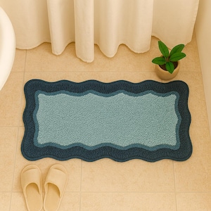 Puede incluir: Una alfombra de baño rectangular con un borde ondulado en tonos azules. La alfombra tiene una superficie texturizada y está rodeada por un ribete azul oscuro. Dos zapatillas beige están colocadas cerca de la alfombra. También se ve una pequeña planta en maceta.