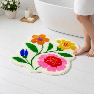 May include: A cream-colored floral bath mat with a whimsical design. The mat features orange, yellow, pink, and blue flowers with green stems and leaves. The mat is placed on a white wooden floor.
