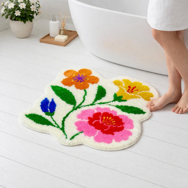 May include: A cream-colored floral bath mat with a whimsical design. The mat features orange, yellow, pink, and blue flowers with green stems and leaves. The mat is placed on a white wooden floor.