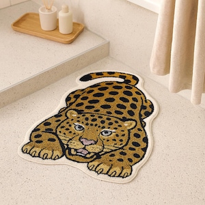 May include: A beige and brown leopard-shaped bath mat with black spots. The mat has a cream-colored border and features a detailed face with blue eyes and a pink tongue. The mat is placed on a light-colored surface.