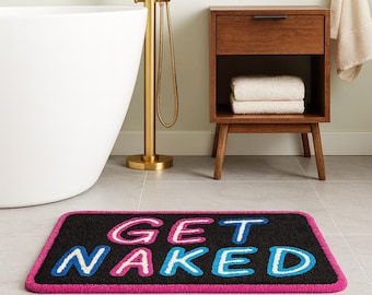 May include: A rectangular black bath mat with the words "GET NAKED" in pink, white, and blue lettering. The mat has a pink border and is placed on a tiled floor near a white bathtub and wooden cabinet.