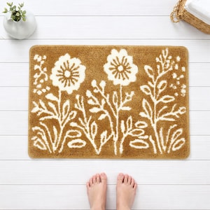 Könnte beinhalten: Eine rechteckige Badematte mit floralem Design. Die Matte hat eine warme braune Farbe mit weißen Blumen- und Blättermustern. Die Matte liegt auf einem weißen Holzboden. Das Bild zeigt auch eine kleine weiße Vase mit einer Pflanze und einen Korb mit weißen Handtüchern.