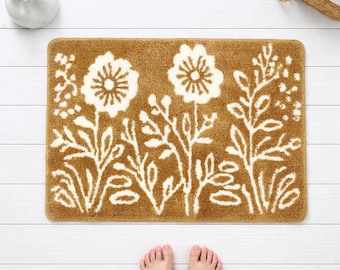 May include: A rectangular bath mat with a floral design. The mat is a warm brown color with white floral and leaf patterns. The mat is on a white wooden floor. The image also shows a small white vase with a plant and a basket with white towels.