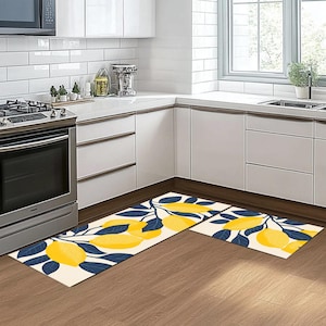 May include: Two kitchen rugs featuring a lemon and leaf design. The rugs are white with blue leaves and yellow lemons. The rugs are positioned in a kitchen with white cabinets and a wooden floor.