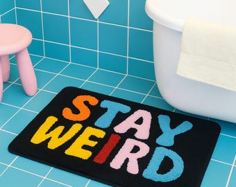 Puede incluir: Alfombra de baño rectangular negra con las palabras "STAY WEIRD" en letras coloridas y texturizadas. La alfombra está en un baño con un taburete rosa, una bañera blanca y baldosas azules.
