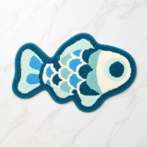 Op de afbeelding: Een visvormige badmat in blauwe, turquoise en witte tinten. De mat heeft een donkerblauwe omtrek, een groot oog en schubachtige patronen. Deze decoratieve mat voegt een speelse touch toe aan elke badkamer. Ongeveer 70 cm lang.