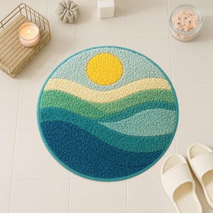 Op de afbeelding: Een ronde badmat met een landschapsontwerp. Het heeft een gele zon, een lichtblauwe lucht en gelaagde groene en blauwe golven. De mat heeft een donkerblauwe rand en ligt op witte tegels. De diameter is ongeveer 60 cm.