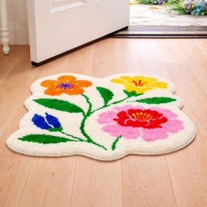 May include: A floral-shaped rug with a cream-colored background. The rug features colorful flowers in orange, yellow, pink, and blue, with green leaves and stems. The rug is placed on a wooden floor near an open door.