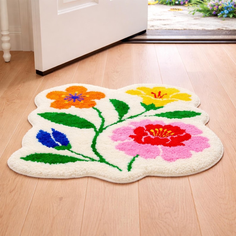 May include: A floral-shaped rug with a cream-colored background. The rug features colorful flowers in orange, yellow, pink, and blue, with green leaves and stems. The rug is placed on a wooden floor near an open door.