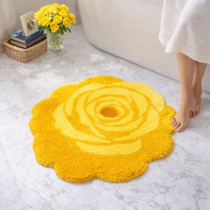 May include: A round, yellow bath mat shaped like a blooming rose with textured, tufted pile. The design features concentric petal layers in varying shades of yellow, creating a floral pattern that covers the entire surface of the mat.