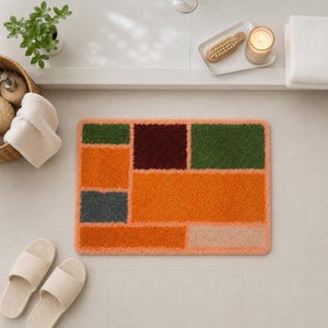 Op de afbeelding: Een rechthoekige badmat met een geometrisch patroon van pluche, getufte blokken in tinten oranje, donkergroen, diepbordeaux, antracietgrijs en beige. De mat heeft een zacht, getextureerd oppervlak met afgeronde hoeken en een lichtoranje rand.