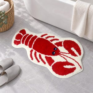 May include: A plush, lobster-shaped bath mat featuring a vibrant red crustacean design with dark blue eyes, set against a cream-colored background. The mat has a soft, tufted texture and follows the contoured silhouette of a lobster with claws.