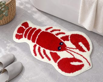 May include: A plush, lobster-shaped bath mat featuring a vibrant red crustacean design with dark blue eyes, set against a cream-colored background. The mat has a soft, tufted texture and follows the contoured silhouette of a lobster with claws.