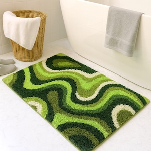 Può includere: Un tappetino da bagno a fantasia verde e bianco con un design ondulato. Il tappeto è realizzato in un materiale morbido e felpato ed è posizionato su un pavimento piastrellato bianco. Un cestino di vimini con un asciugamano bianco e una vasca da bagno sono sullo sfondo.