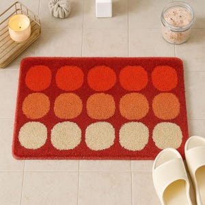 Op de afbeelding: Een rechthoekige badmat met een rode achtergrond en grote, cirkelvormige patronen in tinten oranje en beige. De mat is gemaakt van een zacht, getextureerd materiaal en is ontworpen voor gebruik in de badkamer.