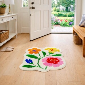 May include: A floral-shaped rug with a cream-colored background and colorful flowers. The flowers are orange, yellow, pink, and blue, with green stems and leaves. The rug is placed on a light wood floor near a white door.
