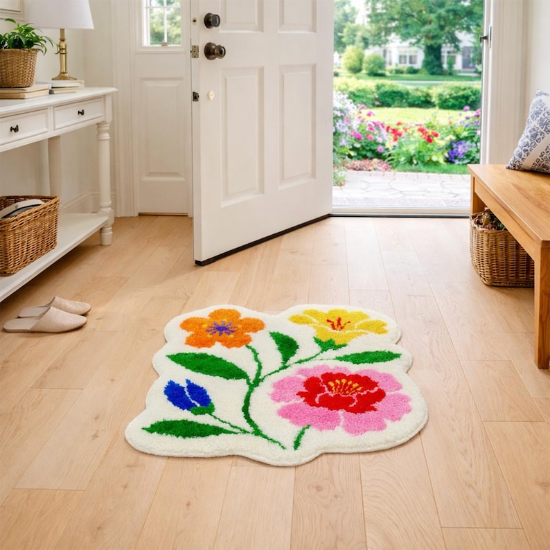 May include: A floral-shaped rug with a cream-colored background and colorful flowers. The flowers are orange, yellow, pink, and blue, with green stems and leaves. The rug is placed on a light wood floor near a white door.