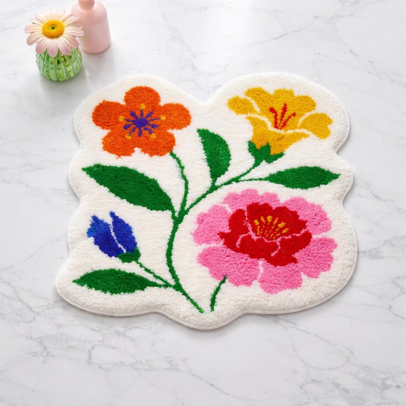 May include: A floral-shaped bath mat with a white background and colorful flowers. The flowers are orange, yellow, pink, and blue, with green stems and leaves. The mat is made of a soft, textured material.