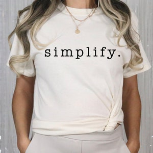 Puede incluir: Una camiseta blanca con la palabra "simplificar" impresa en negro en la parte delantera. La camiseta está anudada a la cintura y la modelo la lleva con pantalones blancos.