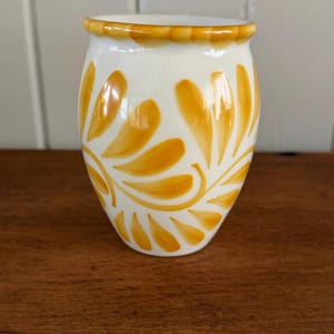 May include: A white ceramic vase with a rounded body and a flared rim, featuring hand-painted golden-yellow leaf patterns that wrap around the exterior. The design consists of stylized, brush-stroked foliage motifs in a warm, vibrant yellow hue.