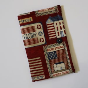 May include: A rectangular fabric wallet in deep red featuring a patriotic print with houses, American flags, and text including BRIGHT STARS, GLORY, BROAD STRIPES, and OL FLAG. The wallet is secured with a single dark circular snap button closure.