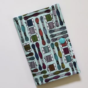May include: A rectangular fabric pouch in light blue featuring a repeating pattern of colorful embroidery floss skeins and bobbins with the word FLOSS printed on them. The pouch is secured with a single, solid light blue plastic snap button.