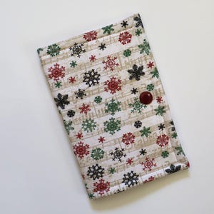 May include: A rectangular fabric case featuring a cream background with a printed pattern of musical notes and scattered snowflakes in red, green, and black. The case is secured with a single dark red circular button on the front flap.