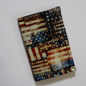 May include: A rectangular fabric wallet featuring a distressed American flag print in red, white, and blue. The wallet is secured with a single dark circular snap button closure, showcasing a weathered, vintage-style pattern of stars and stripes.