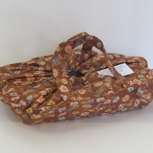 May include: A brown fabric carrier with a floral pattern in blue, orange, and yellow. The carrier has a handle and a tag with text. The fabric is gathered and sewn to create a basket shape.