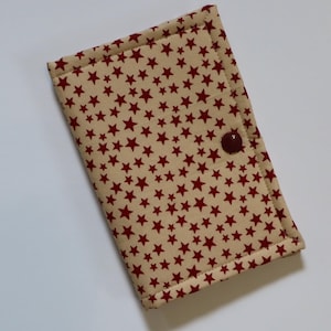 May include: A rectangular fabric wallet or case featuring a beige background with a repeating pattern of small dark red stars. The item is quilted with visible stitching along the edges and includes a single dark red snap button closure.