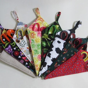 May include: A collection of colorful fabric scissor holders, each with a unique pattern and a pair of scissors inside. Patterns include floral, fruit, cow print, and novelty designs. The scissors have various handle colors.