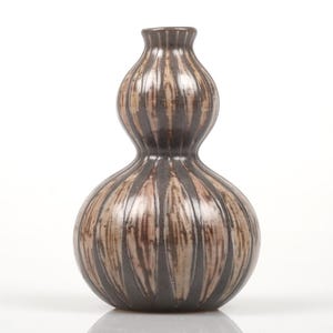 Puede incluir: Un jarrón de cerámica marrón y negro con forma de calabaza y detalles de rayas verticales. El jarrón tiene una base bulbosa y un cuello más estrecho y alargado. La superficie tiene un acabado brillante y el jarrón está sobre un fondo blanco.