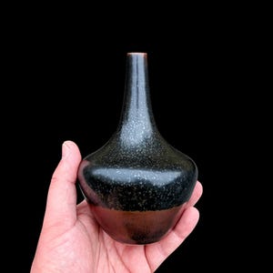 Pode incluir: Um pequeno vaso de cerâmica com pescoço estreito e corpo bulboso. A parte superior apresenta um esmalte preto escuro com uma textura clara salpicada, enquanto a seção inferior transita para um acabamento castanho escuro sólido na base.