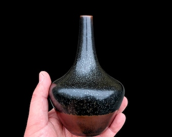 May include: A small ceramic vase with a narrow neck and a bulbous body. The upper portion features a dark black glaze with a speckled, light-colored texture, while the lower section transitions into a smooth, solid dark brown finish at the base.