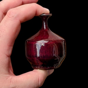 Puede incluir: Un pequeño jarrón de cristal rojo oscuro con cuello estrecho y cuerpo bulboso. El jarrón se sostiene sobre un fondo negro, mostrando su acabado brillante y forma única. El jarrón mide unos 7.6 cm de alto.