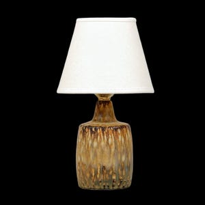 K&ouml;nnte beinhalten: Eine kleine Tischlampe mit wei&szlig;em Lampenschirm und Keramikfu&szlig;. Der Fu&szlig; hat eine braune und goldene Glasur mit vertikalen Tropfenmustern. Die Lampe ist f&uuml;r den Innenbereich konzipiert.