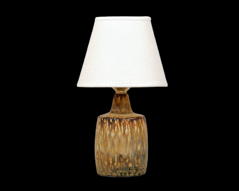 K&ouml;nnte beinhalten: Eine kleine Tischlampe mit wei&szlig;em Lampenschirm und Keramikfu&szlig;. Der Fu&szlig; hat eine braune und goldene Glasur mit vertikalen Tropfenmustern. Die Lampe ist f&uuml;r den Innenbereich konzipiert.