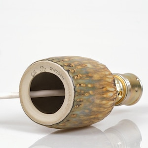 K&ouml;nnte beinhalten: Ein keramischer Lampensockel mit einer strukturierten, gesprenkelten Oberfl&auml;che in Braun-, Gr&uuml;n- und Goldt&ouml;nen. Der Sockel hat eine zylindrische Form mit einer Metallfassung. Der Text "G.N. Sweden" ist sichtbar.