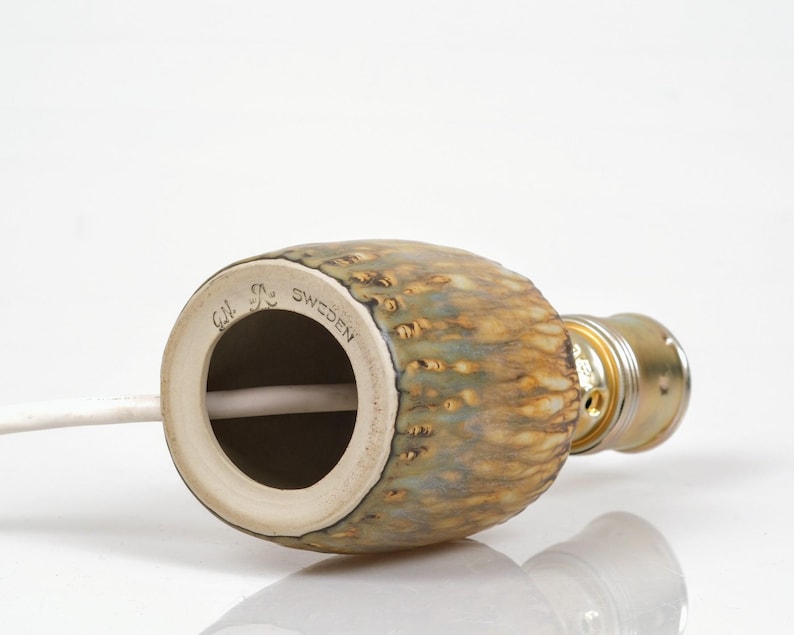 K&ouml;nnte beinhalten: Ein keramischer Lampensockel mit einer strukturierten, gesprenkelten Oberfl&auml;che in Braun-, Gr&uuml;n- und Goldt&ouml;nen. Der Sockel hat eine zylindrische Form mit einer Metallfassung. Der Text "G.N. Sweden" ist sichtbar.
