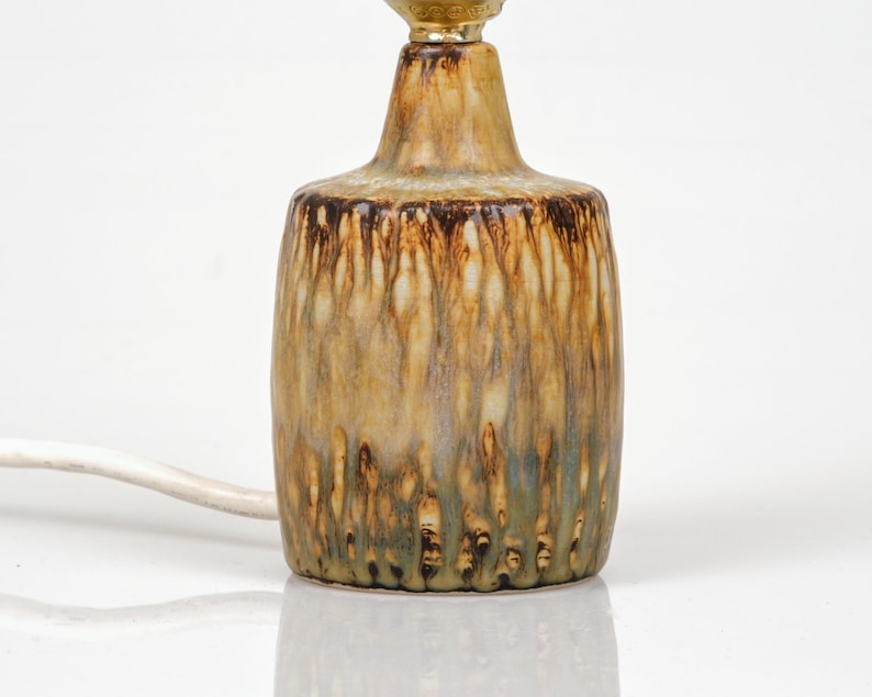 K&ouml;nnte beinhalten: Eine Tischlampe aus Keramik mit strukturierter, glasierter Oberfl&auml;che. Die Lampe hat ein braunes, beiges und gr&uuml;nes Farbschema mit einer goldfarbenen Oberseite. Ein wei&szlig;es Stromkabel erstreckt sich von der Basis.