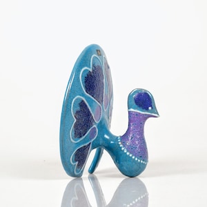Puede incluir: Figurilla de pavo real de cerámica en tonos azules y morados. El pavo real tiene una gran cola en forma de abanico con patrones en forma de corazón. El cuerpo es azul claro con cuello y cabeza morados. La figurilla está sobre una superficie blanca.