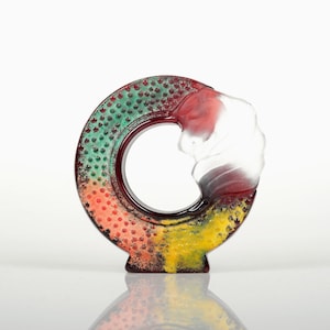 Könnte beinhalten: Eine farbenfrohe, dekorative Glasskulptur in Kreisform. Das Stück weist einen Farbverlauf aus Grün, Rot und Gelb auf, mit einem klaren, wellenartigen Design auf einer Seite. Die Skulptur hat eine strukturierte Oberfläche mit kleinen, gepunkteten Mustern.