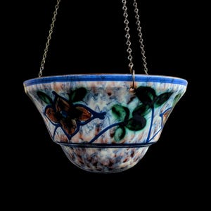 Könnte beinhalten: Ein hängender Keramik-Blumentopf mit floralem Design, aufgehängt an einer silbernen Kette. Der Topf hat einen blauen Rand und ist mit braunen, grünen und blauen Blumenmustern verziert. Der Hintergrund ist schwarz. Durchmesser ca. 20 cm.