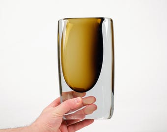 Puede incluir: Un jarrón de vidrio transparente con un interior redondeado de color ámbar. El jarrón tiene forma rectangular con esquinas redondeadas. El color ámbar se concentra en el centro, creando un efecto degradado. El jarrón es sostenido por una mano.