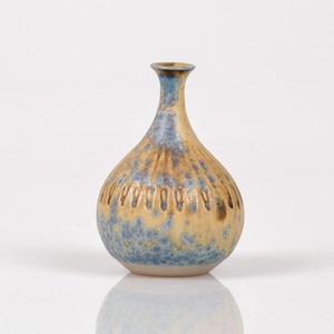 May include: A small, decorative ceramic vase with a bulbous body and a narrow neck. The vase features a textured surface with a pattern of vertical lines and a color gradient of blue, tan, and brown. The vase is approximately 6 inches tall.