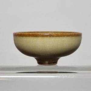 May include: A small, light brown ceramic bowl with a slightly flared rim and a rounded base. The bowl has a subtle glaze that gives it a soft, matte finish.