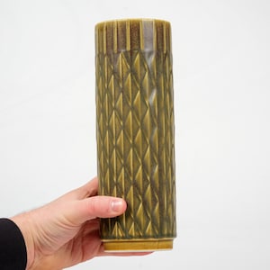 Könnte beinhalten: Eine hohe, zylindrische Vase mit einem geometrischen Rautenmuster in Olivgrün- und Brauntönen. Die Vase wird von einer Hand vor einem weißen Hintergrund gehalten. Die Vase hat eine strukturierte Oberfläche und einen rechteckigen Sockel.