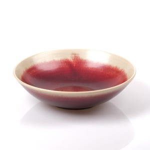 Puede incluir: Un cuenco de cerámica con un diseño ancho y poco profundo. El interior es de un color burdeos intenso que se desvanece en un borde de color crema. El cuenco está sobre un fondo blanco.