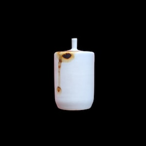 May include: A cylindrical white ceramic vessel with a narrow neck and a flat base. The surface features a distinct brown, rust-colored drip pattern that extends vertically from the top rim down the side, creating a singular organic mark.
